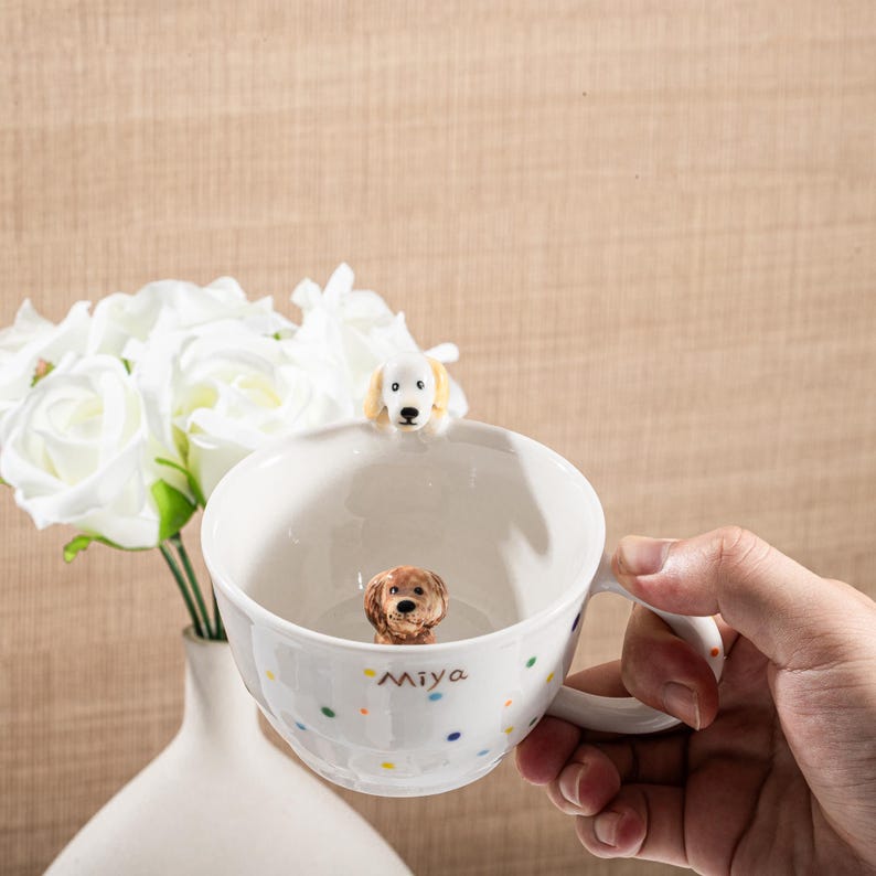 May include: White ceramic mug with a dog figurine inside and on the rim. The mug has a handle and is decorated with colorful polka dots and the name "Miya". A bouquet of white flowers is in the background.
