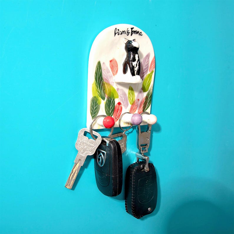 May include: A white ceramic key holder with a black and white cat figurine, the words "Liam & Emma" are written above the cat. The key holder has colorful leaf designs and holds keys and key fobs.