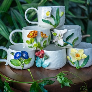 May include: A stack of six white ceramic mugs and cups, each adorned with colorful, three-dimensional floral designs. The flowers include orange, yellow, blue, and red blooms with green leaves. The mugs have handles, and the cups are handle-less.