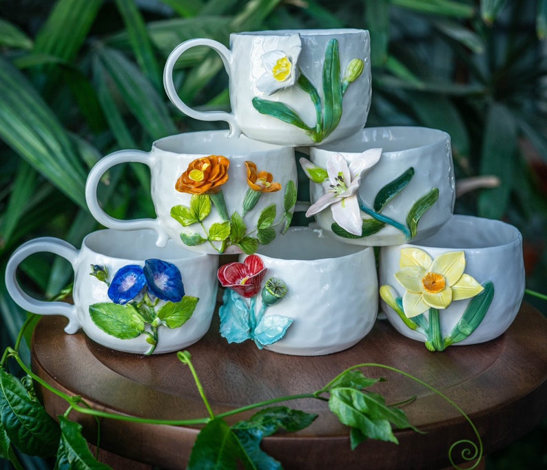 May include: A stack of six white ceramic mugs and cups, each adorned with colorful, three-dimensional floral designs. The flowers include orange, yellow, blue, and red blooms with green leaves. The mugs have handles, and the cups are handle-less.