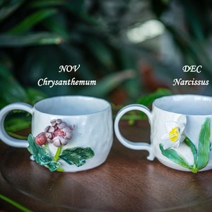 May include: Two white ceramic mugs with floral designs. One mug features a chrysanthemum, the other a narcissus. The text "NOV Chrysanthemum" and "DEC Narcissus" are above each mug. The mugs have a textured surface and a curved handle.
