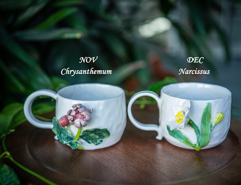 May include: Two white ceramic mugs with floral designs. One mug features a chrysanthemum, the other a narcissus. The text "NOV Chrysanthemum" and "DEC Narcissus" are above each mug. The mugs have a textured surface and a curved handle.