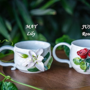 May include: Two white ceramic mugs, one with a lily and the text "MAY Lily", the other with roses and the text "JUN Rose". The mugs are on a wooden tray, with a green leafy background.