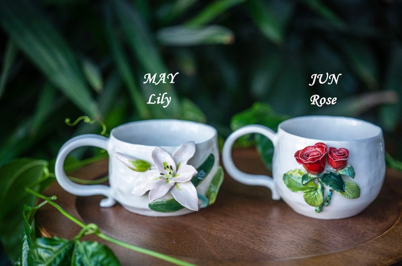 May include: Two white ceramic mugs, one with a lily and the text "MAY Lily", the other with roses and the text "JUN Rose". The mugs are on a wooden tray, with a green leafy background.