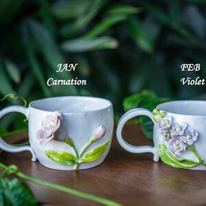 May include: Two white ceramic mugs with floral designs. One mug features a carnation, the other violets. The text "JAN Carnation" and "FEB Violet" are above the mugs. The mugs have curved handles and are set against a blurred green background.