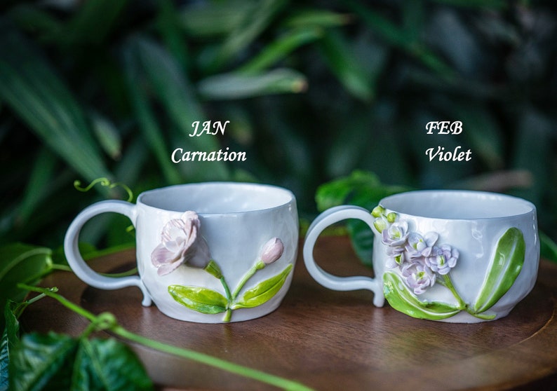 May include: Two white ceramic mugs with floral designs. One mug features a carnation, the other violets. The text "JAN Carnation" and "FEB Violet" are above the mugs. The mugs have curved handles and are set against a blurred green background.