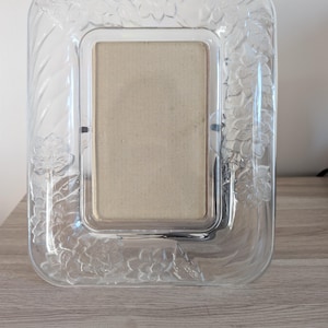Peut inclure: Un cadre photo rectangulaire en verre transparent présentant un motif en relief moulé de fleurs et de feuilles sur tout le pourtour. Le cadre possède des coins arrondis et une ouverture centrale rectangulaire contenant un insert beige uni.