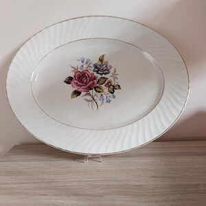 May include: An oval white ceramic serving platter featuring a central floral bouquet with a prominent pink rose, blue bell-shaped flowers, and green leaves. The rim has a textured, wavy, embossed pattern and a thin gold-coloured metallic edge.