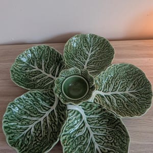 May include: A green ceramic serving platter shaped like a large cabbage leaf with five distinct petal-like sections surrounding a central circular bowl. The surface features a detailed, embossed texture mimicking the veins of a cabbage leaf.