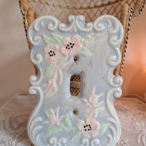 Peut inclure: Plaque d'interrupteur en céramique bleu clair avec un motif floral. La plaque présente des fleurs roses, des feuilles vertes et des volutes blanches. Elle comporte deux trous de vis et une ouverture unique pour l'interrupteur.
