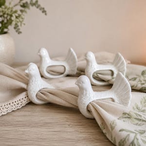 Peut inclure: Un ensemble de quatre ronds de serviette en céramique blanche, chacun en forme d'oiseau stylisé avec une aile et une queue texturées. Les ronds sont disposés sur une serviette en lin beige à motif de feuilles vertes, révélant leur finition brillante.