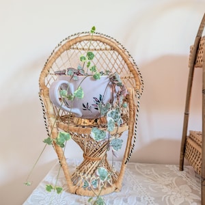 Peut inclure: Une tasse en céramique violet clair avec une base dorée et des motifs floraux est placée dans une petite chaise paon en rotin tressé. Une plante retombante de type chaîne de cœurs aux petites feuilles vertes et blanches en cascade.