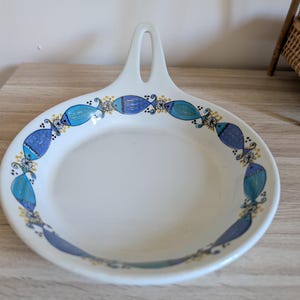 Peut inclure: Un repose-cuillère en céramique blanche avec une poignée et une bordure décorative de poissons bleus et turquoise. Les poissons sont entrecoupés d'accents floraux jaunes et blancs. Le repose-cuillère est sur une surface en bois.