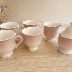 Peut inclure: Un service à thé de six pièces de couleur rose clair, comprenant quatre tasses avec anses, une crémière et un sucrier avec couvercle. L'ensemble présente une base blanche et un effet dégradé, passant du rose au blanc.