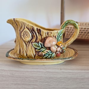 Peut inclure: Une saucière en céramique avec soucoupe intégrée, présentant un motif texturé d'écorce d'arbre dans des tons marron et jaune. L'extérieur est orné de champignons sculptés en relief, de feuilles vertes et de petits glands près de l'anse.