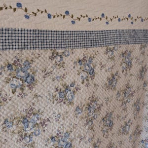 Peut inclure: Un tissu matelassé présentant une section supérieure sur fond blanc avec une vigne horizontale de petites fleurs bleues. En dessous se trouve une bordure à carreaux bleus et blancs, suivie d'une large section au motif floral bleu et violet.