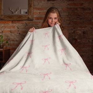 May include: A white blanket with a pink bow pattern. The blanket is being held up by a person.
