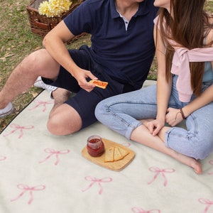 May include: A couple sits on a white picnic blanket with pink bows printed on it. They are enjoying a snack of crackers and jam. The picnic blanket is spread out on a grassy lawn.
