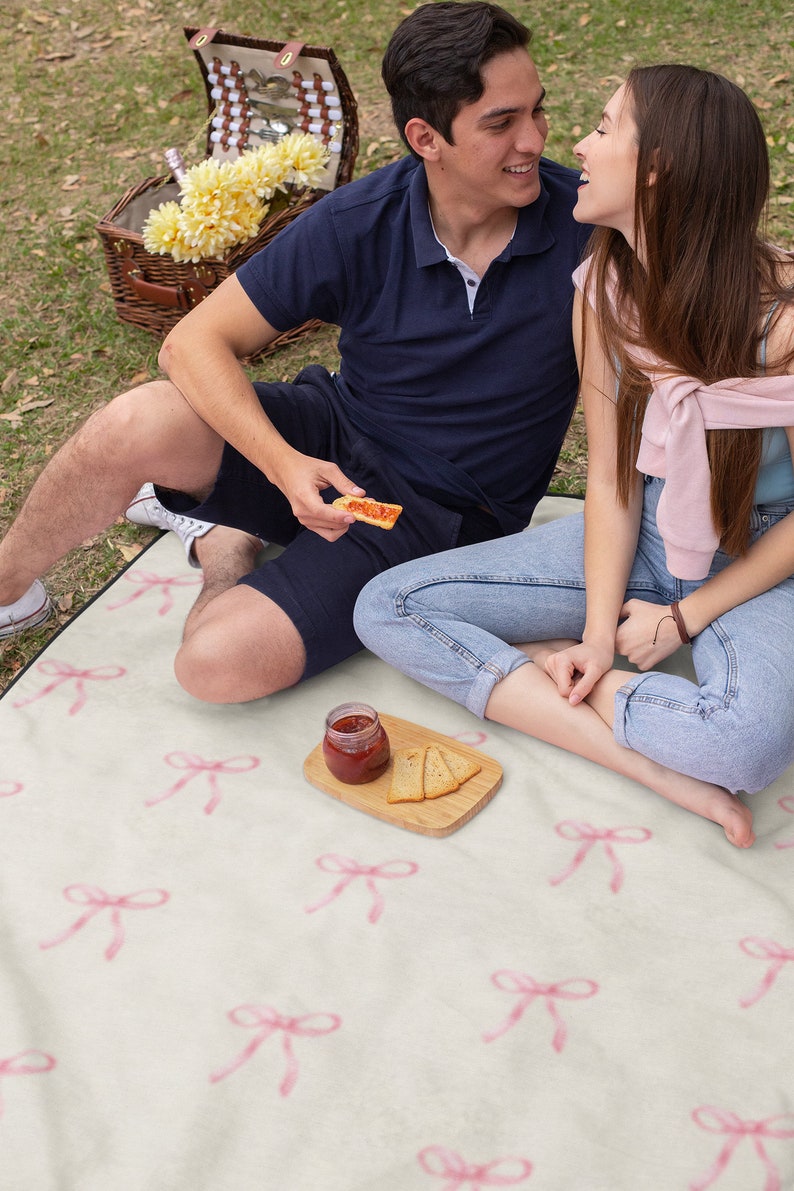 May include: A couple sits on a white picnic blanket with pink bows printed on it. They are enjoying a snack of crackers and jam. The picnic blanket is spread out on a grassy lawn.