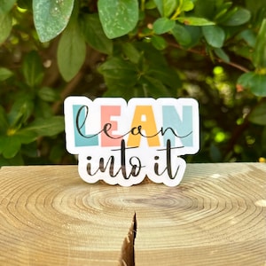 May include: A sticker with the text "lean into it" in colorful letters on a white background. The sticker is on a wooden surface.