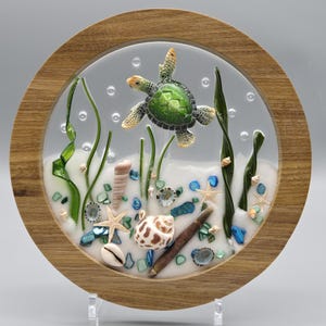 May include: A round wooden framed art piece featuring a sea turtle swimming above a sandy ocean floor. The scene includes seashells, starfish, and green seaweed. The turtle has a green shell and tan flippers.