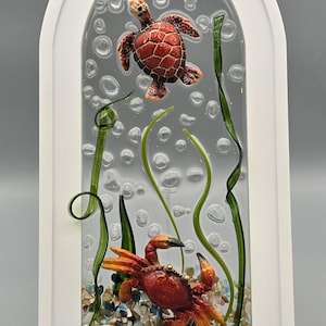 May include: A framed art piece featuring a red sea turtle and crab in a clear resin scene with bubbles, green seaweed, and a sandy seabed. The artwork is encased in a white, arched frame, creating a unique underwater diorama.