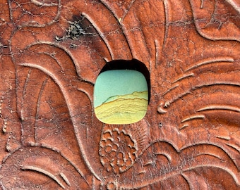 May include: A square, polished stone with rounded corners. The stone features a landscape design with a light blue sky and layered yellow and brown hills. The stone is set against a textured, brown leather background.