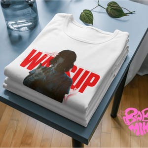 May include: A stack of white t-shirts with a graphic design featuring the text "WASSUP" in red, along with a silhouette of a person. The shirts are neatly folded on a light blue table, with a glass and plant in the background.
