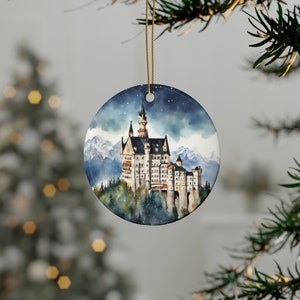 May include: Round ceramic Christmas ornament featuring a watercolor painting of a castle with a mountain backdrop. The castle is white with a dark roof and is set against a blue sky with falling snow. A gold string is attached for hanging.