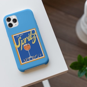 May include: A light blue phone case featuring a graphic with the text "Do you Spritz Aperol?" in yellow and white. The graphic includes a cocktail glass with orange slices. The case is on a white surface.