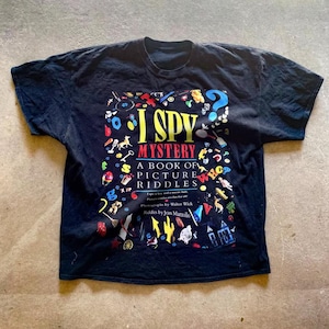 May include: Black t-shirt with a colorful graphic of the book "I Spy Mystery: A Book of Picture Riddles". The graphic features a variety of objects, including a key, a toy car, a house, and a question mark.