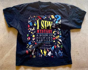 May include: Black t-shirt with a colorful graphic of the book "I Spy Mystery: A Book of Picture Riddles". The graphic features a variety of objects, including a key, a toy car, a house, and a question mark.
