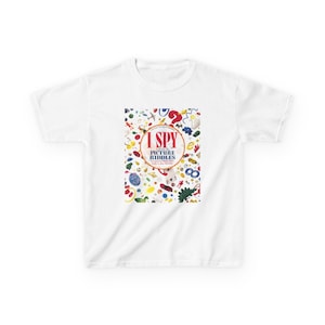 May include: White t-shirt featuring the text "I SPY PICTURE RIDDLES" in a red and gold circular design. The shirt's graphic includes various colorful objects, creating a playful and engaging visual for children's apparel.