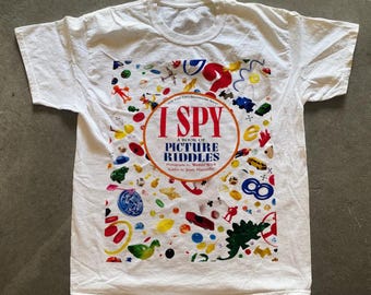 May include: White t-shirt with a colorful print of the book cover "I Spy: A Book of Picture Riddles" by Walter Wick. The print features a variety of objects, including toys, animals, and household items.