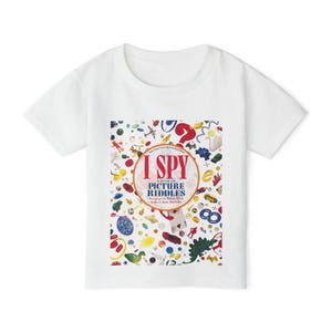 May include: A white short-sleeved t-shirt featuring a central graphic print of the I Spy A Book of Picture Riddles cover. The design includes the red and blue text I Spy A Book of Picture Riddles surrounded by various colorful small objects.