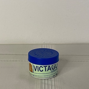 Op de afbeelding: Een kleine, ronde pot met een blauwe deksel en een witte basis. Op de pot staat het woord "VICTAGU" in vette, zwarte letters. Het etiket bevat ook de woorden "Fabriqué en France".