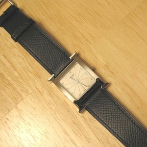 May include: A silver square-shaped wristwatch with a white face and black leather band. The watch has a unique design with a double-loop strap.