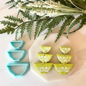 Pu&ograve; includere: Un paio di orecchini statement verdi e bianchi con un design a ciotola impilata. Gli orecchini sono realizzati in pasta polimerica e presentano un motivo floreale bianco.