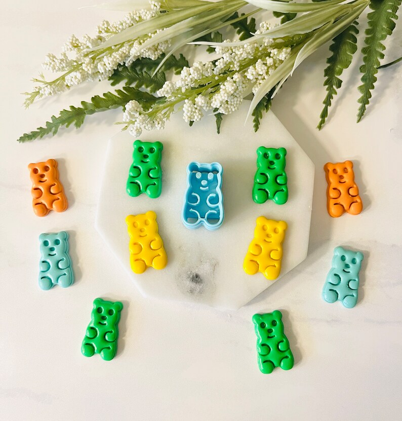 May include: A set of gummy bear cookie cutters in various colours, including green, blue, yellow, and orange. The cutters are arranged on a white surface with a white flower sprig in the background.