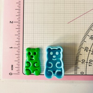 May include: Two gummy bear cookie cutters, one green and one blue. The cutters are on a white gridded surface with a pink ruler and protractor.