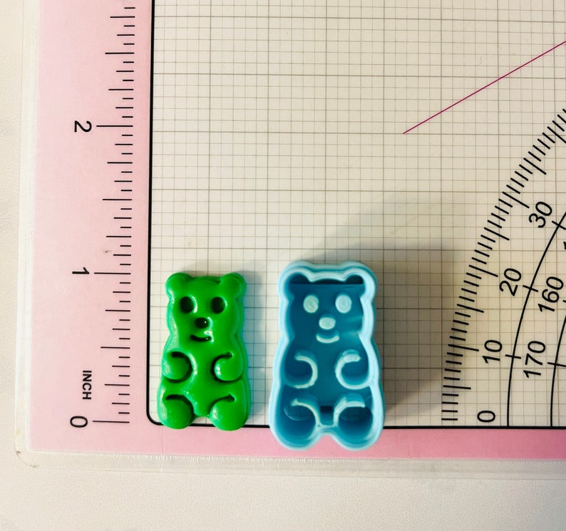 May include: Two gummy bear cookie cutters, one green and one blue. The cutters are on a white gridded surface with a pink ruler and protractor.