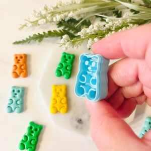 May include: A set of gummy bear moulds in various colours, including green, blue, yellow, and orange. The moulds are made of plastic and are shaped like gummy bears. A hand is holding a blue gummy bear mould.