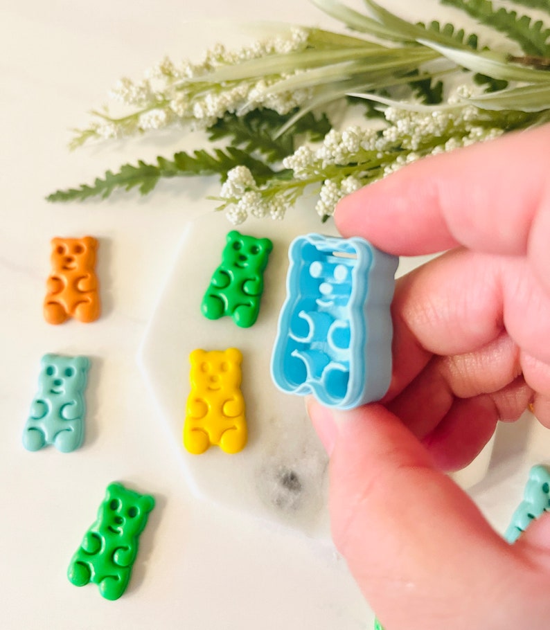 May include: A set of gummy bear moulds in various colours, including green, blue, yellow, and orange. The moulds are made of plastic and are shaped like gummy bears. A hand is holding a blue gummy bear mould.