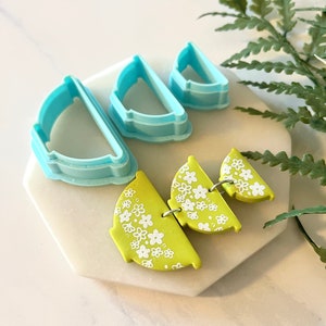 Pu&ograve; includere: Tre tagliabiscotti in plastica blu a forma di semicerchi, con un tagliabiscotti pi&ugrave; piccolo annidato all'interno di un tagliabiscotti pi&ugrave; grande. Un set di tre orecchini in argilla polimerica verde lime con un motivo floreale bianco.
