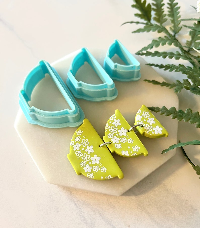 Pu&ograve; includere: Tre tagliabiscotti in plastica blu a forma di semicerchi, con un tagliabiscotti pi&ugrave; piccolo annidato all'interno di un tagliabiscotti pi&ugrave; grande. Un set di tre orecchini in argilla polimerica verde lime con un motivo floreale bianco.