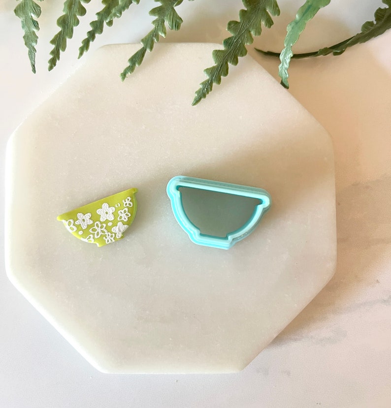 Pu&ograve; includere: Un tagliapasta a forma di ciotola verde e bianca e un tagliapasta a forma di ciotola in plastica blu chiaro su una superficie di marmo bianco.