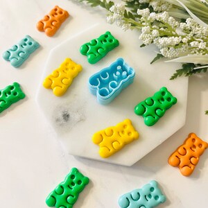 May include: A set of colourful gummy bear cookie cutters in various colours, including green, blue, yellow, and orange. The cutters are arranged on a white surface with a sprig of white flowers in the background.