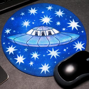 Peut inclure: Tapis de souris rond bleu avec une illustration d'OVNI et trois silhouettes de chats noirs dans les fenêtres, entouré d'étoiles blanches. Une souris d'ordinateur noire et un bloc-notes sont également visibles.