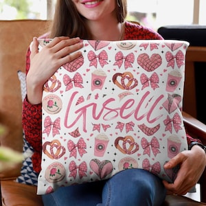 May include: A white cushion with a pink and white pattern of bows, hearts, pretzels, and coffee cups. The name "Giselle" is written in pink cursive lettering on the cushion.