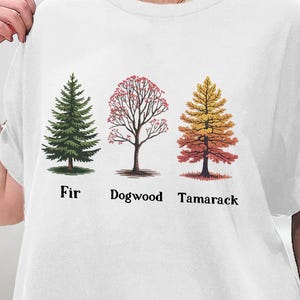 Può includere: Maglietta bianca con una grafica di tre alberi in diversi colori e stadi di crescita. Gli alberi sono etichettati "Fir", "Dogwood" e "Tamarack".